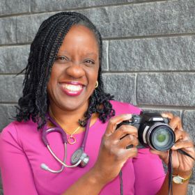 Smiling woman in purple dress with a stethoscope around her neck, holding a camera